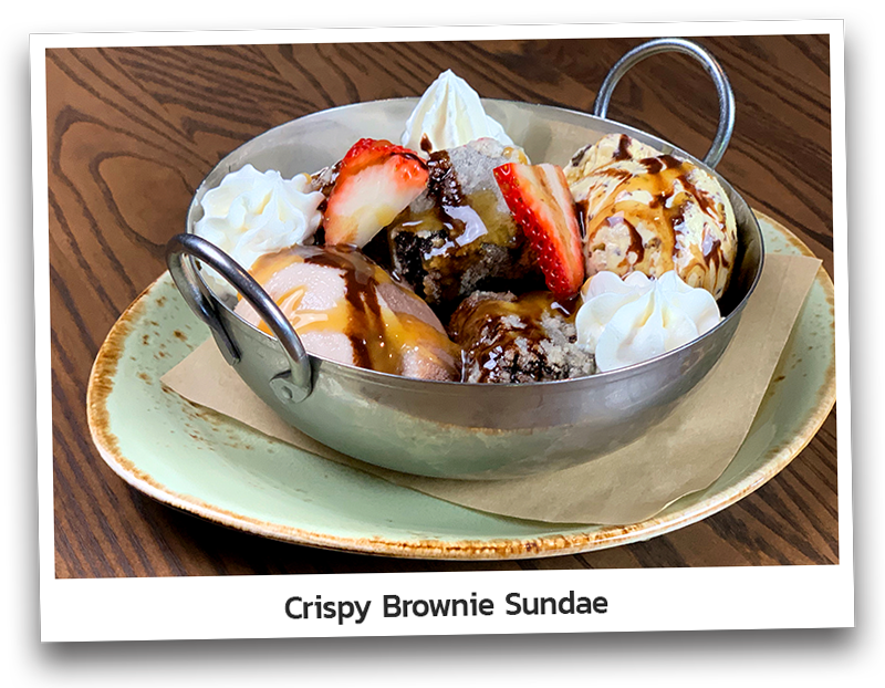Picture of the Crispy Brownie Sundae including bits of crispy brownie layered with vanilla, chocolate and cookies &amp; cream ice cream. Topped with caramel, chocolate syrup, strawberries and whipped cream and presented on a metal bowl.
