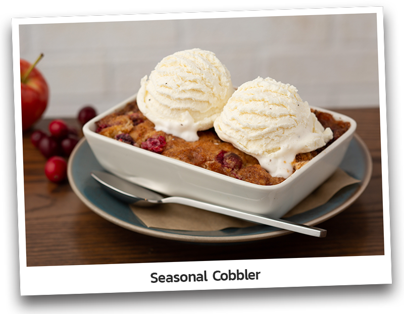 Picture the Seasonal Cobbler which includes seasonal fruit topped with a brown sugar and butter crumble. Served with vanilla ice cream white bowl dish.